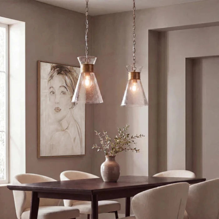 A single pendant light with an antique brass ceiling canopy, chain, and collar, holding a clear textured glass shade and a warm Edison-style bulb, shown against a plain white background.